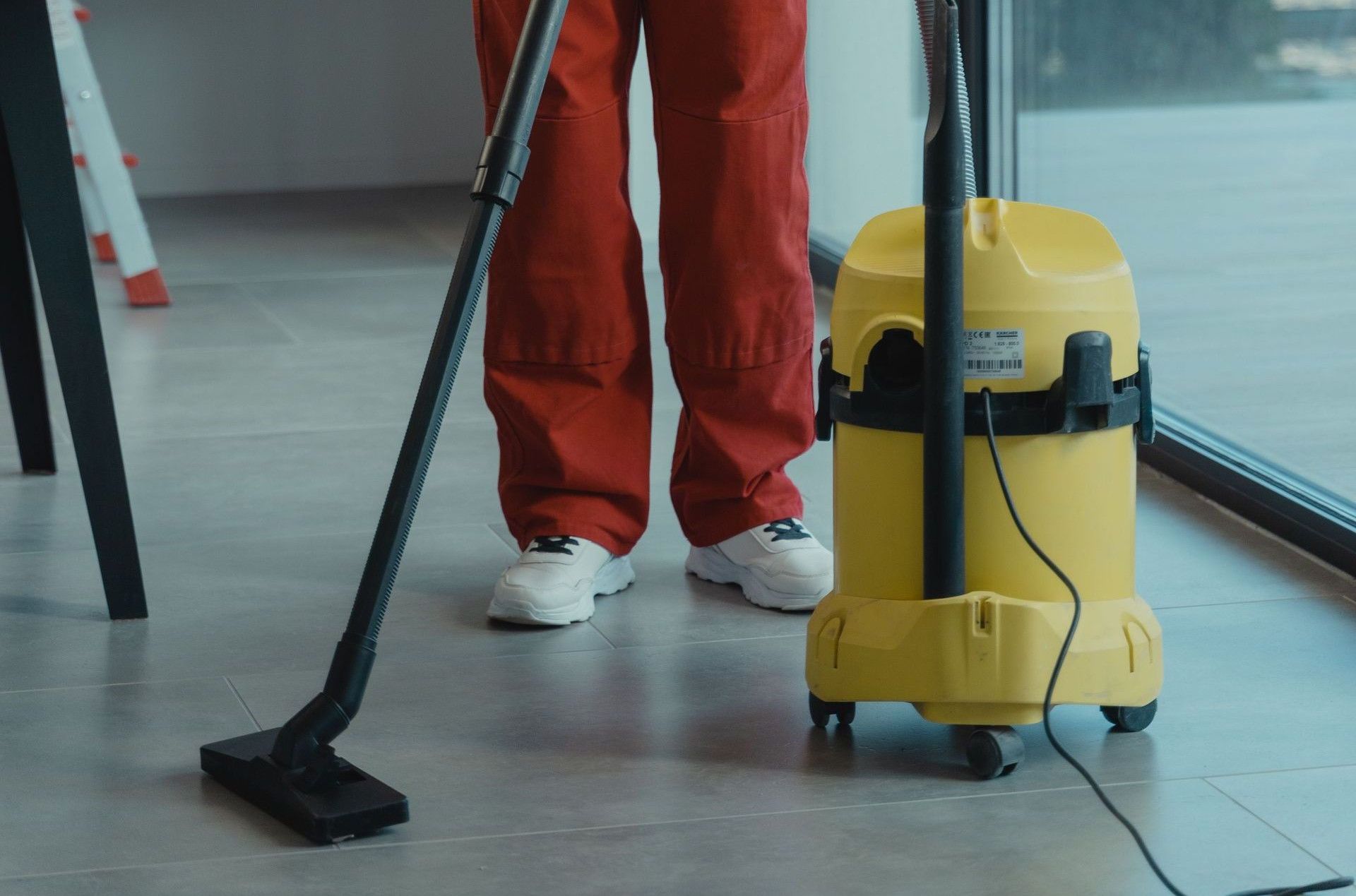An adult woman in red coveralls operates a vacuum cleaner in a modern indoor setting, focusing on cleaning tasks.