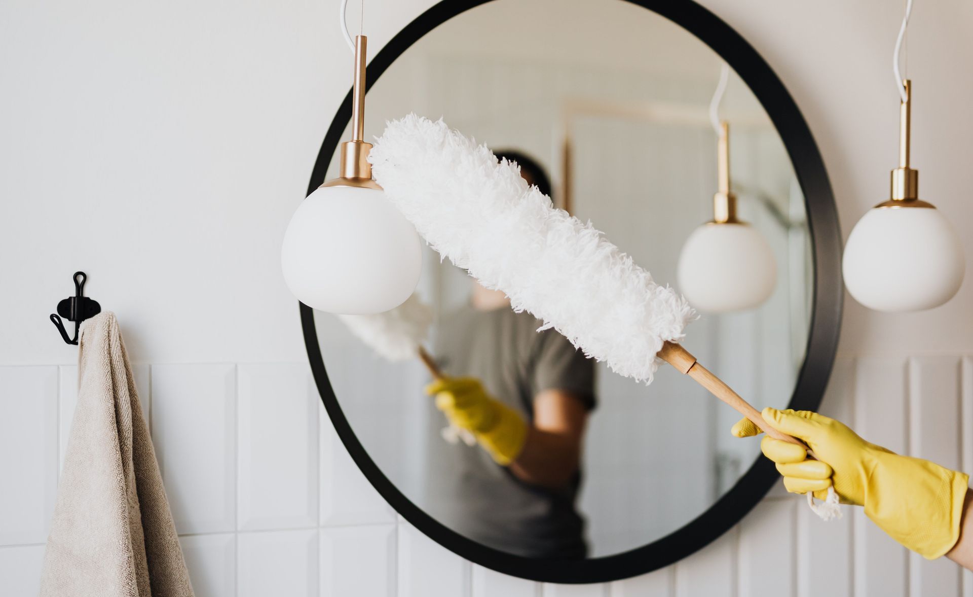 Crop anonymous female housewife in rubber gloves dusting modern lamp with soft brush against mirror while cleaning stylish bathroom